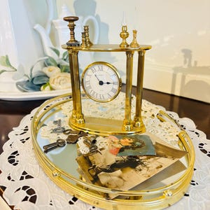 May include: A gold-toned clock with Roman numerals on a mirrored tray with keys and a vintage photograph. The clock is on a decorative stand with two small candle holders.
