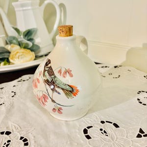 May include: White ceramic pitcher with a cork stopper and a painted design of a bird with pink flowers.