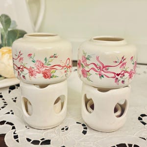 May include: Two cream-colored ceramic candle holders. Each has a cylindrical top decorated with pink roses and red ribbons. The base is a white, openwork design. The candle holders are displayed on a white lace tablecloth.