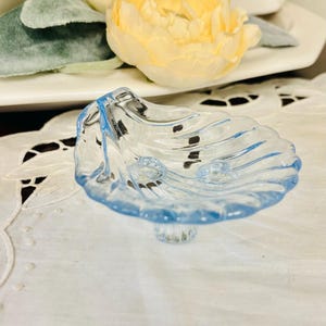 May include: A light blue glass dish shaped like a seashell with a scalloped edge and a small pedestal base. The translucent material features molded ridges that radiate from the center, creating a textured surface that catches the light.