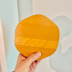 May include: A light-colored, wooden serving board with a unique, organic shape. The board is made of several wooden planks joined together, creating a textured surface. The board is held by a person's hand, showcasing its size and design.
