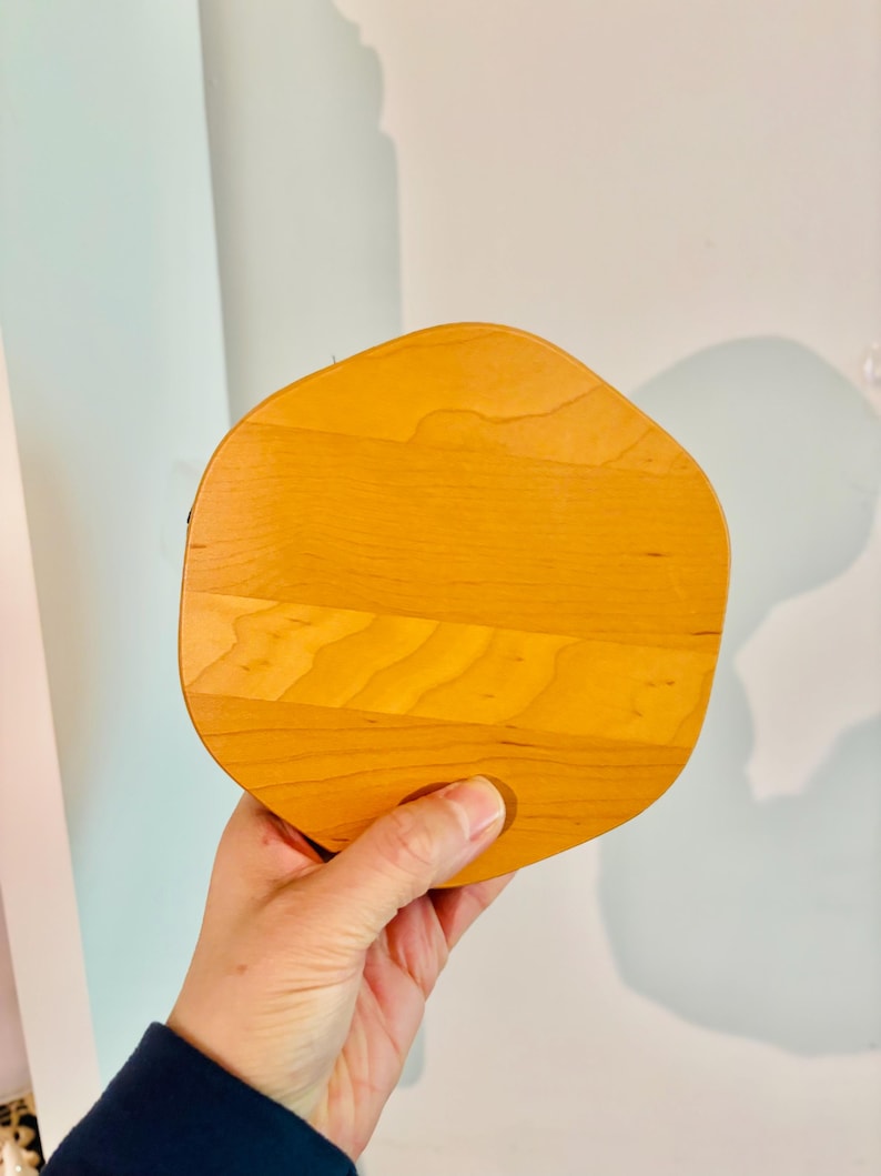 May include: A light-colored, wooden serving board with a unique, organic shape. The board is made of several wooden planks joined together, creating a textured surface. The board is held by a person's hand, showcasing its size and design.