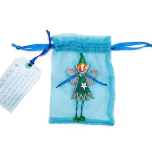 May include: A blue tulle pouch with a drawstring closure. The pouch contains a small felt tooth fairy with green wings, a green dress, and a star on her chest. The pouch has a tag attached that reads "Tooth Pixie" and includes instructions for placing a lost tooth inside.