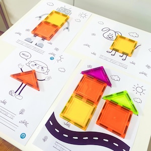 May include: Four sheets of white paper with black line drawings and colourful magnetic tiles. The drawings include a robot, a dog, a girl, and houses. The magnetic tiles are orange, yellow, green, and purple.