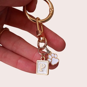 May include: Gold-tone keychain with a large round clasp and a smaller rectangular charm with the letter "P" in white. A white paw print charm hangs next to it. The keychain is held by a hand.