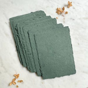 May include: A stack of handmade, teal-colored paper sheets with rough, deckled edges. The paper has a textured appearance and is arranged on a white marble surface, with dried flowers in the background. Ideal for stationery or art projects.