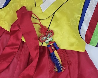 Könnte beinhalten: Ein traditionelles koreanisches Hanbok-Kleid für ein Kind. Das Oberteil ist gelb mit einem Blumenmuster und der Rock ist rot. Das Kleid hat einen dekorativen Knoten und eine Quaste.