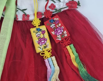 Könnte beinhalten: Zwei traditionelle koreanische Knotenamulette, bekannt als "Jogak", mit gestickten Blumendesigns. Die Amulette sind rot und gelb mit aufwendigen Details und sind an langen, bunten Quasten befestigt.