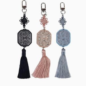 May include: Three decorative keychains with detailed designs. Each keychain has a metal pendant with a detailed pattern, a tassel, and a decorative knot. The keychains are in black, rose gold, and blue-grey colours.