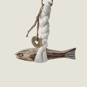 Peut inclure: Ornement en forme de poisson en bois, avec une corde blanche et une pièce de monnaie dorée. Le poisson est brun clair avec des accents bruns et violets plus foncés. La corde est attachée à un anneau doré en haut.
