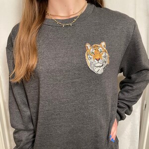 May include: A dark gray long-sleeved sweatshirt with an embroidered tiger face on the left chest. The tiger is orange, black, and white.