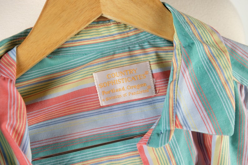 May include: A striped button-down shirt with a light blue, yellow, green, and pink color scheme. The shirt has a label that reads "Country Sophisticates, Portland, Oregon, a division of Pendleton."