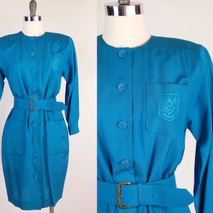 May include: Teal button-down dress with long sleeves and a matching belt. The dress has a small pocket on the chest with an embroidered crest. The dress is displayed on a mannequin.