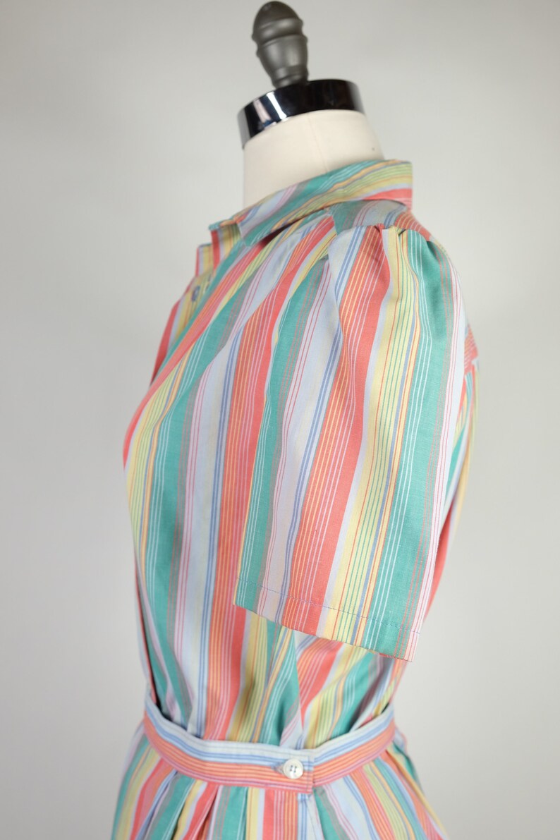 May include: A striped dress with a light blue, pink, green, and yellow color scheme. The dress has a button-up front and a short sleeve with a puff shoulder detail. The dress has a belt.