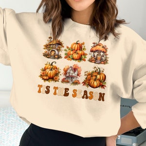 May include: A cream-coloured sweatshirt with a graphic print of pumpkins, houses, and the text "Tis the Season".
