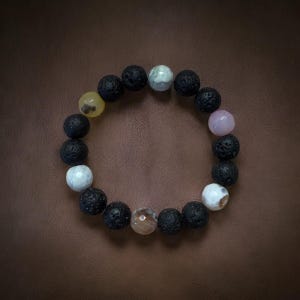 May include: A circular bracelet composed of various stone beads. The bracelet features a mix of black lava rock beads and colorful accent beads in shades of white, yellow, pink, and brown. The bracelet is displayed on a brown surface.