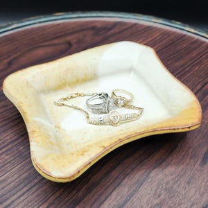 May include: A square, cream-colored ceramic dish with a gold rim holds a collection of jewelry. The jewelry includes a gold bracelet with a heart-shaped pendant, two rings, and a silver ring with a diamond-like stone.