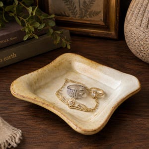 May include: A square, cream-colored ceramic trinket dish with a speckled finish and slightly raised, wavy edges. Inside the dish rests a gold-toned filigree bracelet with a central stone, a silver-toned ring, and a gold-toned ring with a small stone.