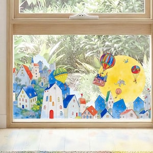 May include: A window decal featuring a colorful illustration of a town with white and blue houses, green trees, and a large yellow moon. Several hot air balloons float above the buildings, with small figures and an elephant depicted in the scene.