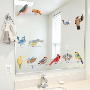 May include: A bathroom mirror decorated with colorful bird decals. The birds are perched on thin, horizontal lines, and feature various colors like red, blue, yellow, and orange. The mirror is above a white countertop with a chrome faucet and a small soap dish.