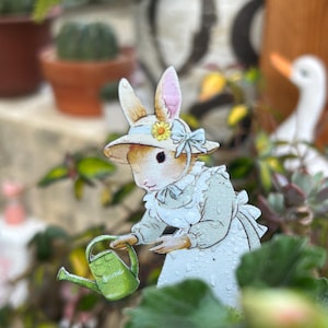 Può includere: Ritaglio di carta di un coniglietto con cappello e vestito, con un annaffiatoio verde. Il coniglietto è circondato da piante e fiori, creando una scena di giardino affascinante. L'abito del coniglietto è azzurro e bianco.