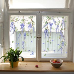 Può includere: Una finestra con un design floreale di fiori di glicine viola e foglie verdi. Il design include farfalle ed è applicato sul vetro. Una piccola pianta in un vaso giallo e una ciotola di frutta sono sul davanzale.