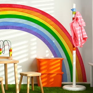 May include: A rainbow wall decal with a colorful, multi-colored design. The rainbow is painted on a white wall. There are two orange storage bins and a white coat rack with a pink shirt hanging on it. The room is decorated with a green artificial grass floor.