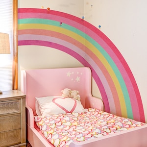 Könnte beinhalten: Ein rosa Kinderbett mit einem Regenbogen-Wandaufkleber und einem Bettwäsche-Set mit Herz-Muster. Das Bett hat ein Kopfteil mit darauf gemalten Sternen.