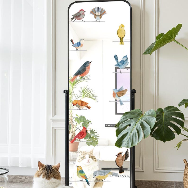 May include: A full-length, black-framed mirror with decorative bird decals. The mirror reflects a room with a cat, plants, and a window. The birds are various colors and perched on small, white shelves.