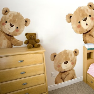 Könnte beinhalten: Eine Szene im Kinderzimmer mit einem kleinen, braunen Teddybär-Spielzeug und drei großen Wandaufklebern mit Cartoon-Teddybären. Die Aufkleber haben rosige Wangen und befinden sich an einer weißen Wand, mit einer Holzkommode und einem Nachttisch.