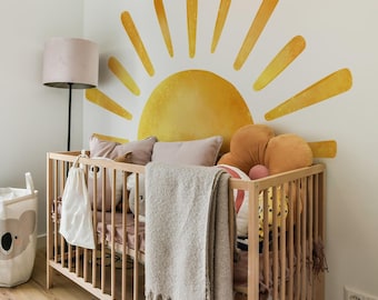 May include: A wooden crib with a white blanket and pillows, decorated with a large yellow sun wall decal. The sun has a watercolor effect and is surrounded by rays.