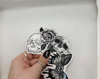 May include: A collection of black and white stickers. The designs include two skulls, one with a rose, and a stylized butterfly. The stickers are held against a white background. The stickers are suitable for various applications.