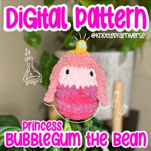 Puede incluir: Un juguete de peluche de ganchillo rosa y blanco de la princesa Chicle del dibujo animado Adventure Time. El juguete lleva una corona dorada con una gema azul. El texto "Princess Bubblegum the Bean" está escrito debajo del juguete.