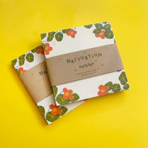 May include: Two square notepads with a floral design of orange flowers and green leaves on a cream background. The notepads are wrapped with a brown paper band that reads "NASTURTIUM Notepad". The background is yellow.