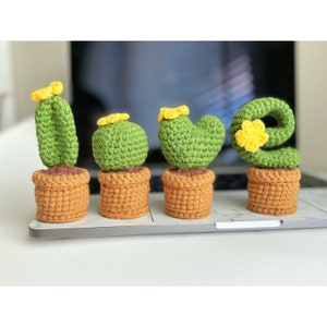 May include: Four crocheted cactus plants in brown pots. The cactus plants are green and have yellow flowers. The cactus plants spell out the word "LOVE".