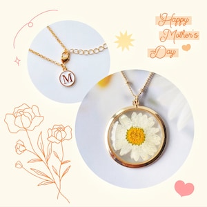 May include: Gold necklace with a round pendant featuring a pressed white daisy with a yellow centre. The pendant is attached to a delicate gold chain. The necklace also includes a separate initial charm with the letter "M" in gold.
