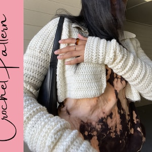 Puede incluir: Un cárdigan blanco de ganchillo recortado con mangas largas. El cárdigan se lleva sobre una camisa de teñido anudado marrón y blanco.