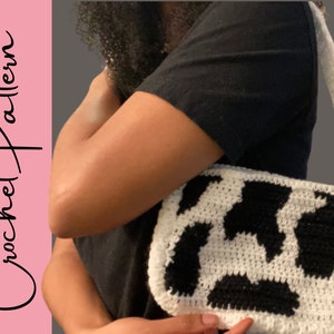 May include: A white and black crocheted handbag with a cow print design. The bag has a long strap and is worn over the shoulder.