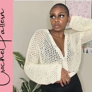 May include: A close-up of a crocheted cardigan with a button closure. The cardigan is off-white and has a loose, open weave. The model is wearing the cardigan over a black top.
