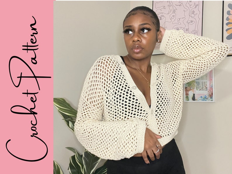 May include: A close-up of a crocheted cardigan with a button closure. The cardigan is off-white and has a loose, open weave. The model is wearing the cardigan over a black top.
