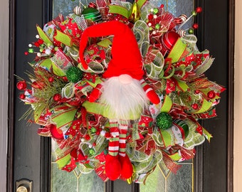 May include: A festive Christmas wreath with a gnome figure. The wreath features red and green ribbons, ornaments, and a plush gnome with a red hat and striped legs. The wreath is hanging on a black door.