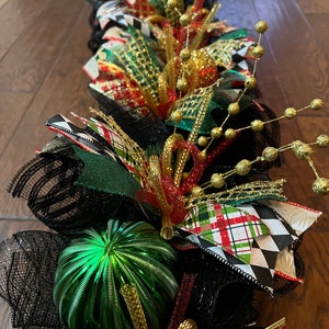May include: A festive Christmas garland featuring a mix of textures and colors. The garland incorporates black mesh, green and red ribbons, gold accents, and a shiny green ornament. The design includes a variety of patterns, including plaid and harlequin.
