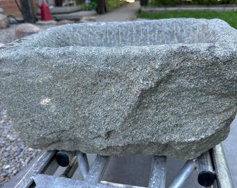 May include: A rectangular stone basin with a rough, textured exterior surface and a deep, hollowed-out center. The stone is a mottled gray and green color with natural variations in the rock face, featuring sharp, uneven edges along the top rim.