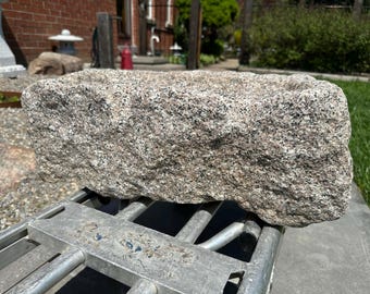 May include: A rectangular stone trough with a rough, chiseled exterior surface. The stone features a speckled pattern of gray, white, and tan mineral grains. The top surface is hollowed out, creating a basin with thick, uneven, and natural edges.