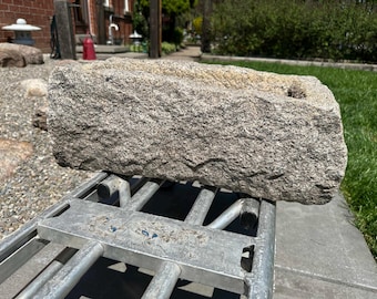May include: A rectangular stone trough with a rough, textured exterior surface in shades of gray and beige. The top of the stone block features a hollowed-out basin with visible chisel marks along the interior edges and a solid, weathered base.