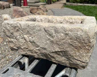 May include: A rectangular stone planter with a rough, chiseled exterior texture and a natural beige and gray color palette. The basin features thick, solid walls and a deep, hollow center, showcasing the raw, weathered appearance of the natural rock.