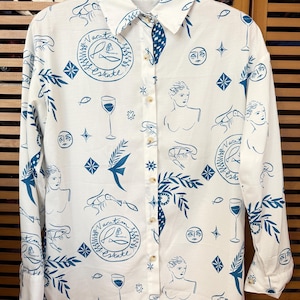 Könnte beinhalten: Weißes Button-Down-Hemd mit blauem Muster mit Illustrationen von Frauen, Fischen, Weingläsern und dem Text "Vacation Estate". Das langärmlige Hemd hat einen Kragen und geknöpfte Manschetten.