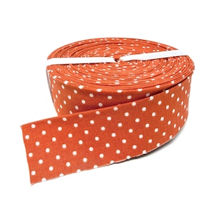 May include: A spool of orange fabric ribbon featuring a repeating pattern of small white polka dots. The ribbon is tightly wound into a circular roll and secured with a thin white band, with one end of the ribbon extending outward from the roll.
