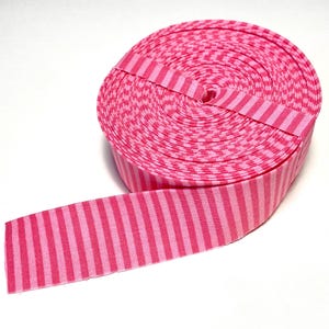May include: A roll of fabric ribbon featuring a repeating pattern of vertical stripes in two distinct shades of pink. The ribbon is tightly wound into a circular spool shape, with one end extending outward to display the consistent striped design.