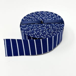 May include: A roll of dark blue ribbon featuring a pattern of thin, vertical white stripes. The ribbon is tightly wound into a circular spool and secured with a thin, solid dark blue band wrapped horizontally around the center of the roll.